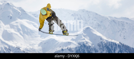 Snowboarder in woollen hat and camouflage trousers performs a tail grab aerial jump with dramatic mountains in the background. Stock Photo