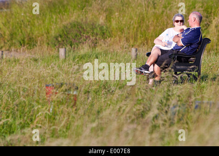 Elderly couple sit on a bench in sunshine surrounded by grass in Brill village, Buckinghamshire Stock Photo