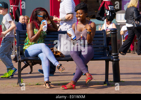 Two black teenage girls sitting on a bench drinking soft drinks. Carlisle Town Centre, Carlisle, Cumbria, England, UK. Stock Photo