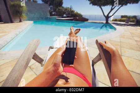 Tanned woman using smart phone by the poolside. Caucasian female model sunbathing on a deckchair and mobile phone. Stock Photo