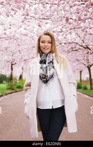 Beautiful young woman wearing long coat standing at spring blossom park. Pretty young caucasian female fashion model posing. Stock Photo