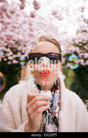 Cute young female wearing sunglasses blowing bubbles at spring park. Beautiful caucasian woman blowing soap bubbles. Stock Photo