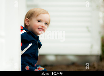 a blond toddler boy sits and smiles on the porch of a white house. Stock Photo