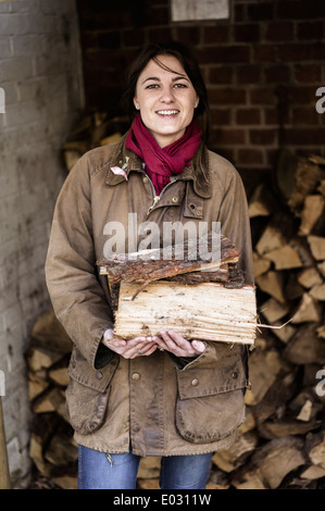 A woman carrying split logs for firewood from a large log store. Stock Photo