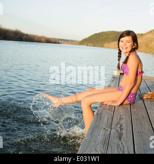 A girl in a bikini sitting on a jetty with her feet in the water. Stock Photo