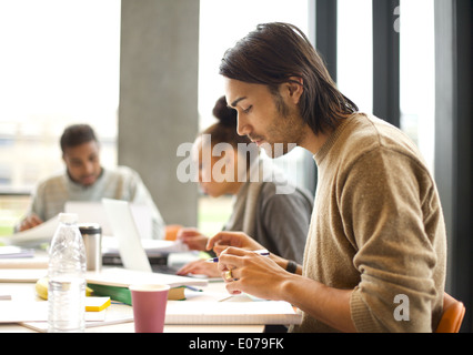 Young man talking notes for study with students studying in background. University students preparing for final exams in library