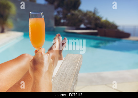 Young woman holding fruit juice while sitting on a deck chair. Female model sunbathing near swimming pool with a glass of juice. Stock Photo