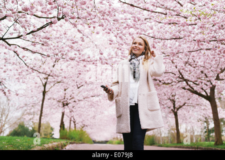 Smiling young woman with mobile phone listening to music in park. Attractive young female at spring blossom garden. Stock Photo