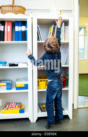 Rear view of boy reaching for container on shelf in kindergarten Stock Photo