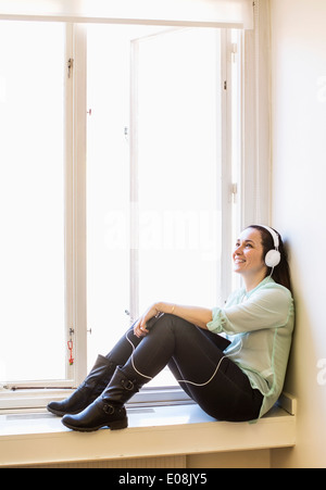 Smiling businesswoman listening music while sitting on window sill Stock Photo