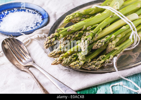 Bunch of young green asparagus on vintage tray served with ceramic plate of sea salt and silver cutlery over green wooden table. Stock Photo