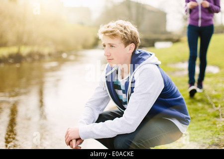 Unhappy teenage boy crouching on riverbank Stock Photo
