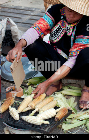 A woman in embroidered ethnic dress roasts corn cobs over charcoal in Yunnan, China Stock Photo
