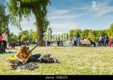 A Young Woman sitting and playing the guitar beside the lake in The Regents Park London England UK Stock Photo