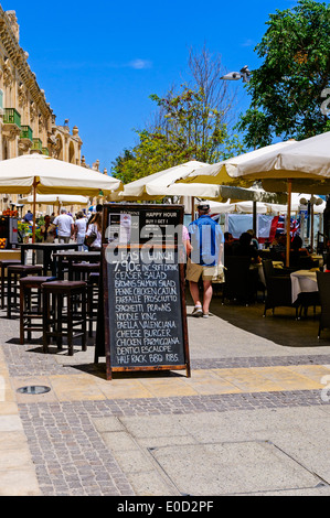 Tourists walk past a menu-board and through a typical restaurant with tables set on either side of the path at the harbour Stock Photo
