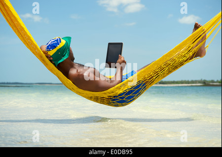 Brazilian man using tablet computer relaxing in hammock on beach over the sea Stock Photo