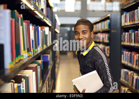 Image of happy young man standing by bookshelf in library. African american student in public library holding books. Stock Photo