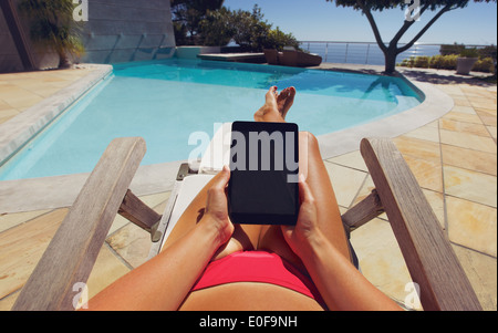 Young woman using a tablet PC near the pool. User POV. Female model relaxing on a deckchair holding digital tablet. Stock Photo