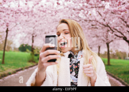 Cheerful young woman talking her picture with her cell phone and gesturing thumbs up sign. Caucasian female model posing. Stock Photo