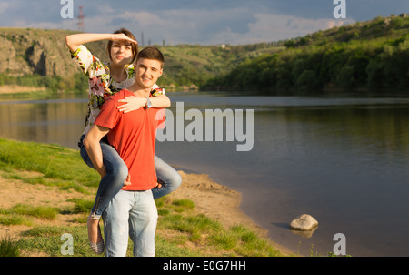 Attractive young couple at a scenic mountain lake enjoying a day in the sunshine with the young man giving his girlfriend a pigg Stock Photo