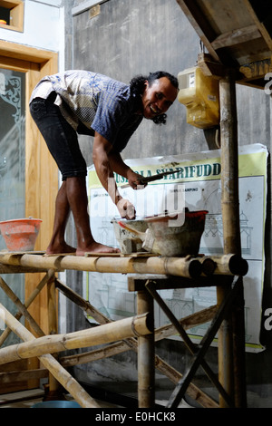 indonesian local villager working at night by the light of a single strip lamp adding plaster and cement to a wall Stock Photo