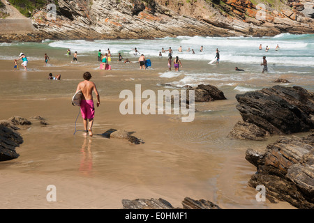 Beach scene with surfer walking on shore with swimmers in background, Victoria Bay Beach, Garden Route, South Africa Stock Photo