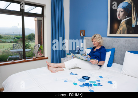 Woman relaxes with a brochure on a white bed at a guesthouse, Victoria Bay, Garden Route, Western Cape, South Africa Stock Photo