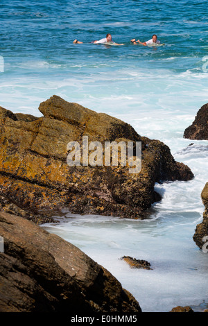 Two male surfers swimming out to sea lying on surfboards in distance from a rocky shore in Victoria Bay, South Africa Stock Photo