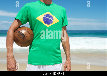 Brazilian soccer player in Brazil flag shirt holding vintage football on tropical Ipanema Beach Rio de Janeiro Stock Photo