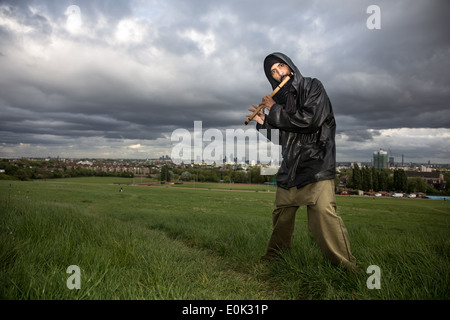mixed race man playing flute in the long grass on hampstead heath wearing headscarf and black shiny jacket Stock Photo