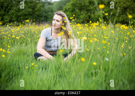 Blonde lady wearing black leggings and a grey crop top sitting in long grass with yellow flowers hampstead heath. Stock Photo