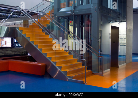 A modern interior featuring a vibrant orange staircase with glass railings alongside a sleek glass elevator. Clean contemporary architectural design. Stock Photo