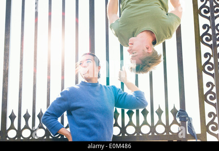 Young man trying to impress indifferent girl by hanging upside down on an iron gate. Stock Photo