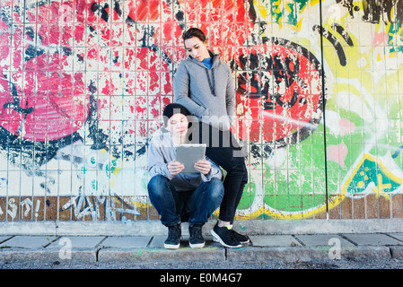 Portrait of casual and relaxed young man and woman reading on a notepad in front of grafitti wall in urban area. Stock Photo