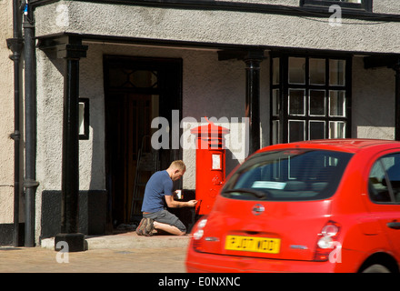 Man painting a pillar box red, as red car passes, Kirkland, Kendal, Cumbria, England UK Stock Photo