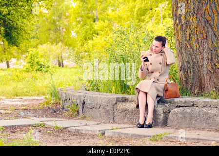 Elegant businesswoman sitting on a stone wall during a sunny spring day, and setting her hair looking herself in a little mirror Stock Photo