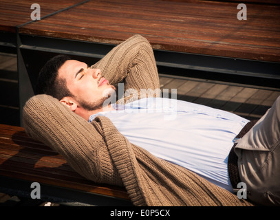 Attractive young man laying down on wood bench in the sun, eyes closed Stock Photo