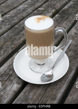 Coffee latte in glass mug on white saucer with spoon on old wooden table England, UK Stock Photo