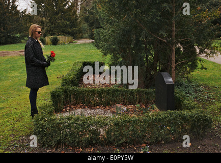 Sad woman grieving in a cemetery holding red roses. Young woman standing at the gravestone of her family member. Stock Photo