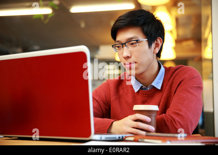 Young asian man in glasses working on laptop and holding cup of coffee Stock Photo