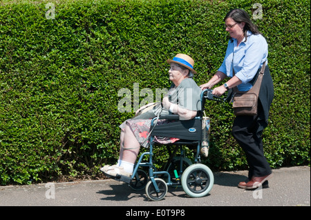 An elderly lady wearing a sun hat is pushed along the pavement in a wheelchair by her carer or assistant on a sunny day. Stock Photo