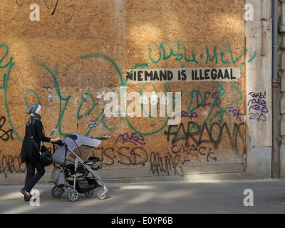 Woman walking pushing pram with graffiti saying nobody is illegal in Dutch language, Brussels, Belgium Stock Photo