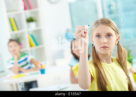 Portrait of lovely girl doing sums on transparent board with schoolmates on background Stock Photo