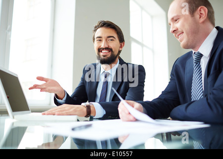 Image of smiling businessman pointing at laptop screen while explaining ideas to his colleague at meeting and looking at camera Stock Photo