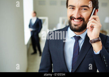 Image of young businessman speaking on the phone and looking at camera on background of his colleague Stock Photo