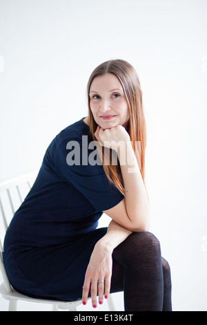 Studio portrait of happy smiling attractive woman wearing blue blouse and sitting in chair and resting chin on hand Stock Photo