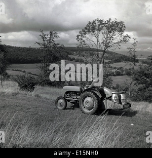 1950s, historical, a farmer driving his tractor across a field with two metal milk churns on the back, with the rolling countryside behind, England. Stock Photo