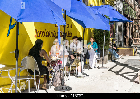 people enjoying Sunday brunch at sidewalk tables under bright blue umbrellas outside Mexican restaurant Hells Kitchen Manhattan Stock Photo