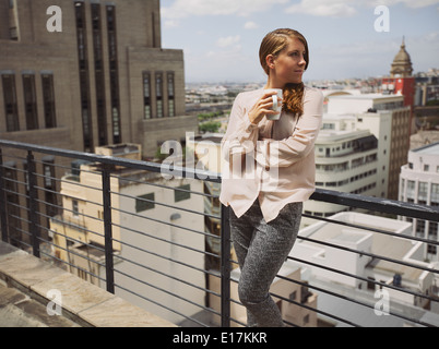 Beautiful young lady standing by a railing on terrace holding a cup of coffee looking away. Caucasian female drinking coffee. Stock Photo