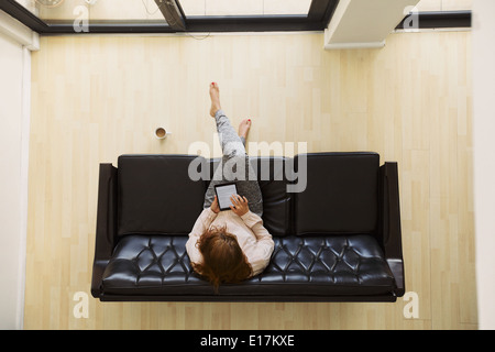 Top view of relaxed young lady sitting on a sofa working on a digital tablet. Aerial view of female sitting on couch with tablet Stock Photo